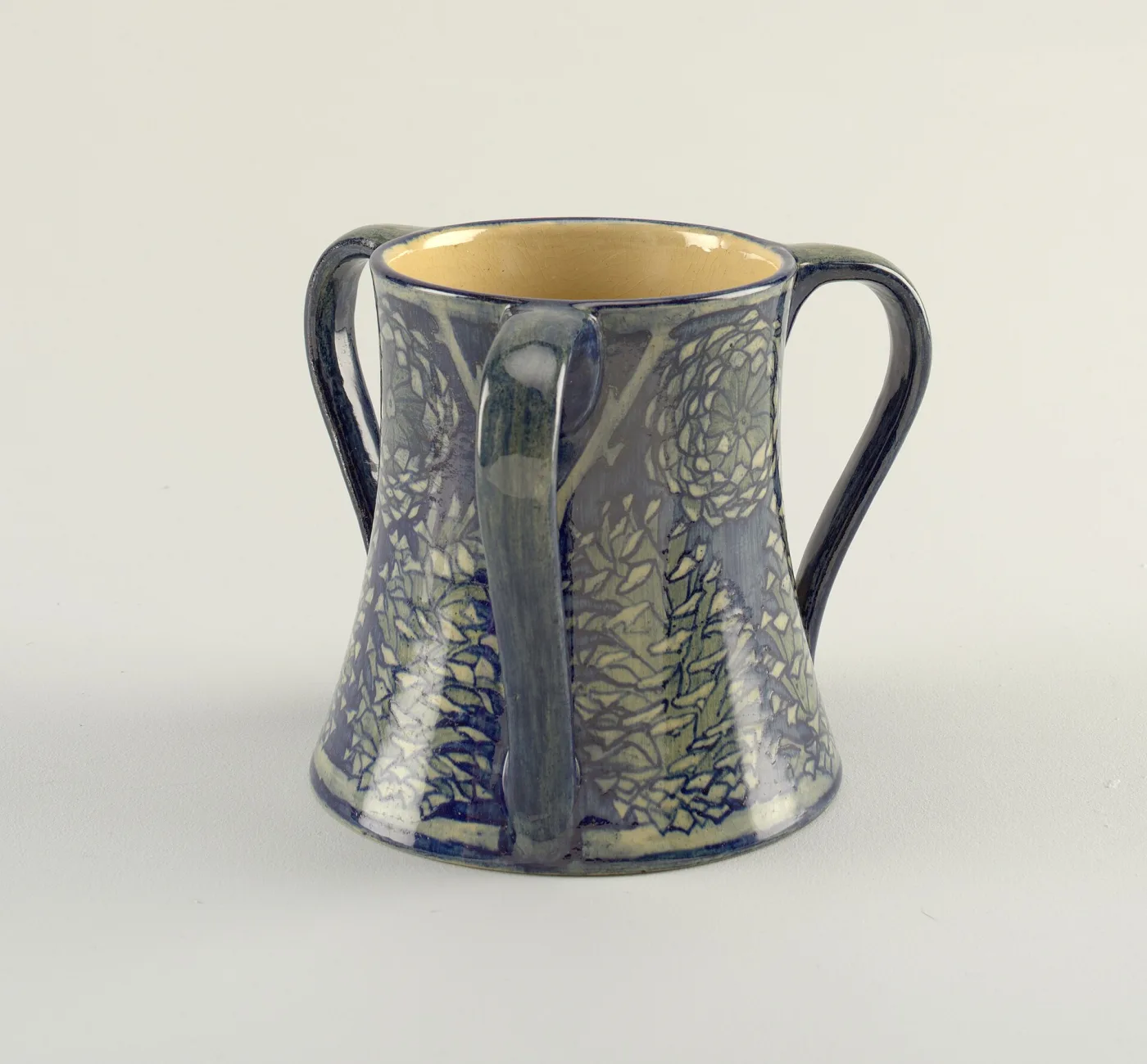 Two-handled tapering mug in soft blue and green matte glaze, carved and painted around the body with a row of trees hung with pale foliage