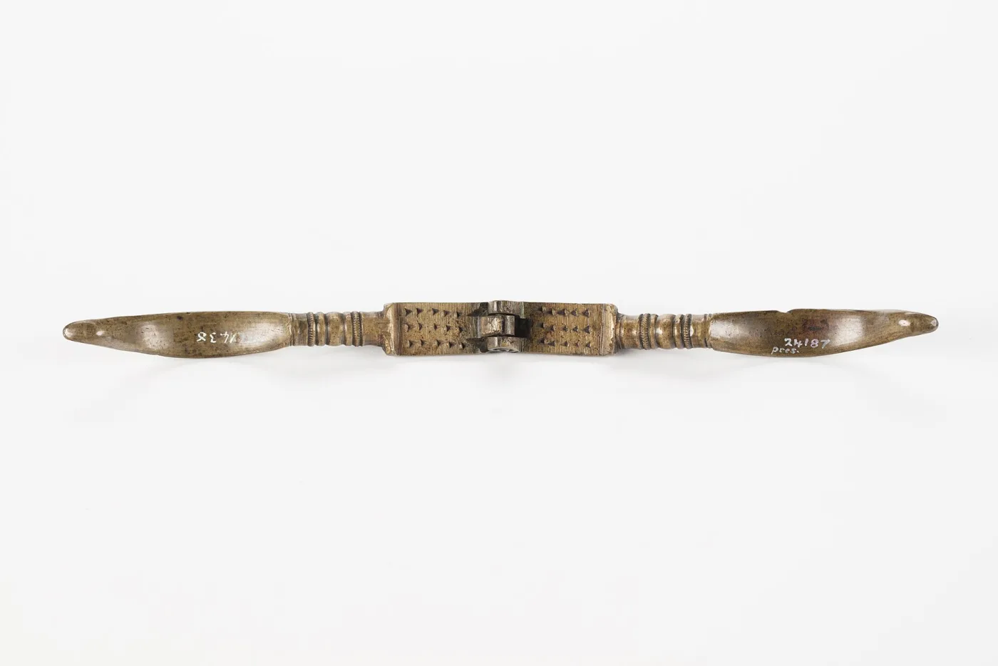 The same brass nutcracker opened out flat, showing the two punched hinge blocks meeting at a single riveted pivot and pale accession numbers painted near each terminal