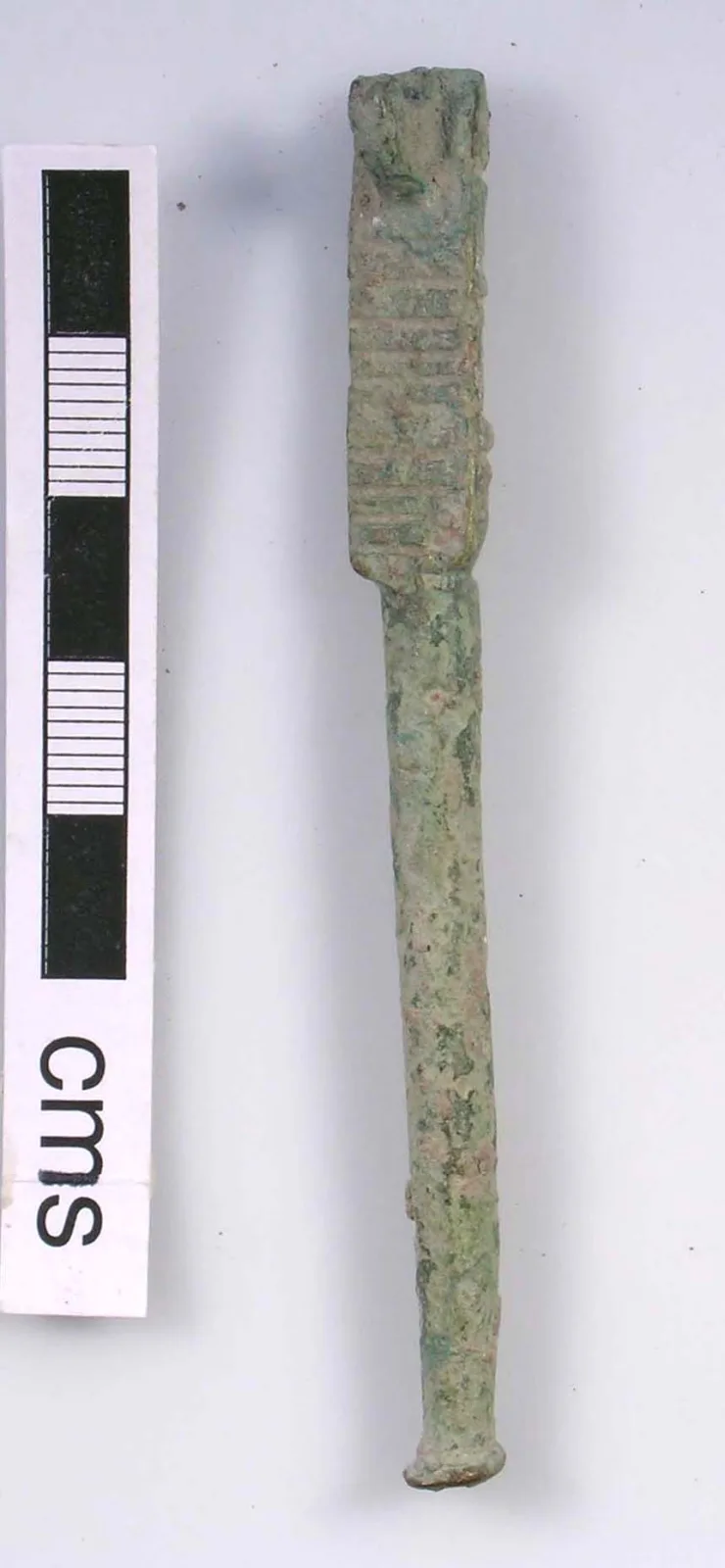 Single corroded copper-alloy nutcracker arm photographed beside a centimetre scale, with a ridged rectangular head above a plain tapering handle