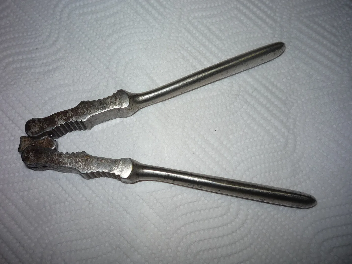 A double-sided lever nutcracker lying on white paper, its long handles bright and its ridged jaws darkened and pitted