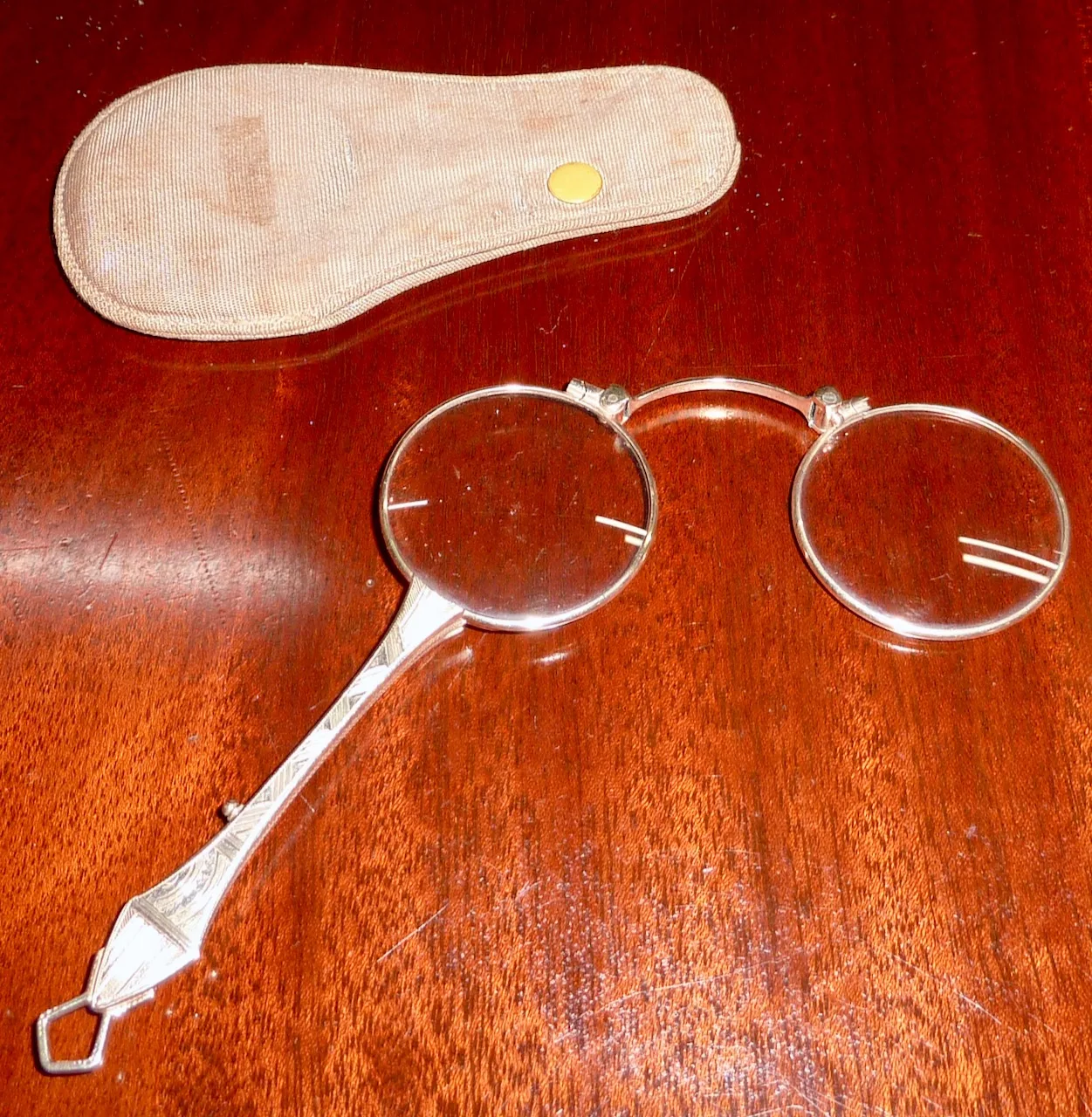 Folding silver-coloured lorgnette spectacles with an engraved handle, open beside a fabric slip case