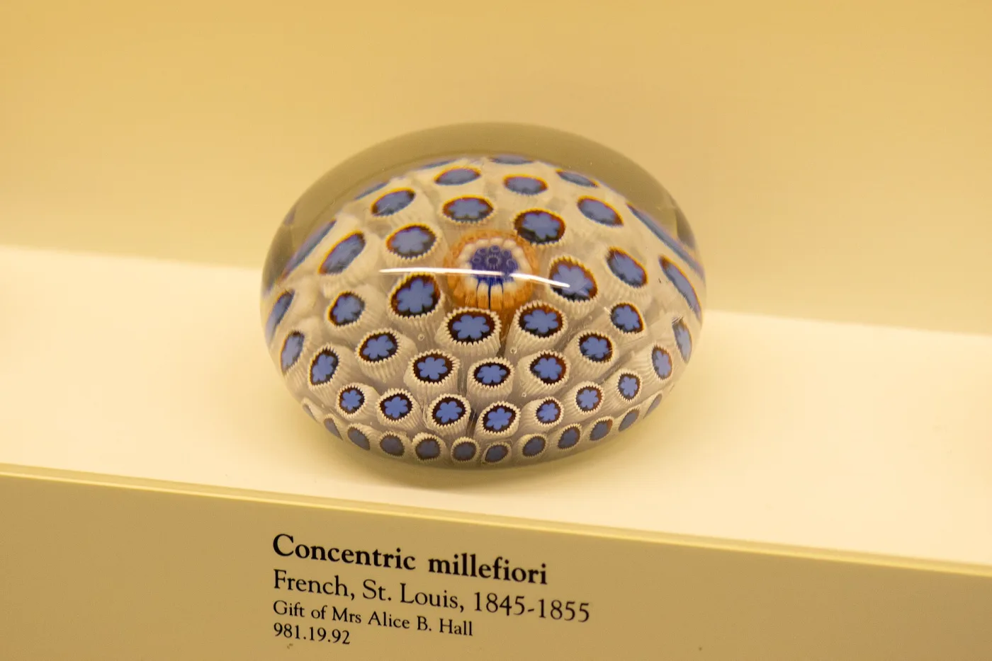 Domed paperweight of concentric millefiori canes in blue, white and amber, resting on a museum shelf beside a printed label reading Concentric millefiori, French, St. Louis, 1845-1855