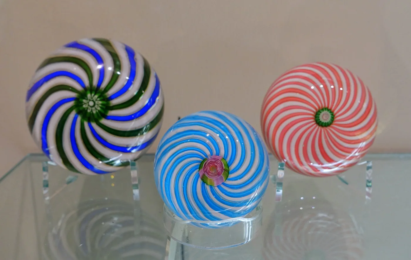 Three swirl paperweights standing in a row on a glass shelf: one green and white, one blue and white, one pink and white, each with alternating opaque spiral ribbons radiating from a central cane