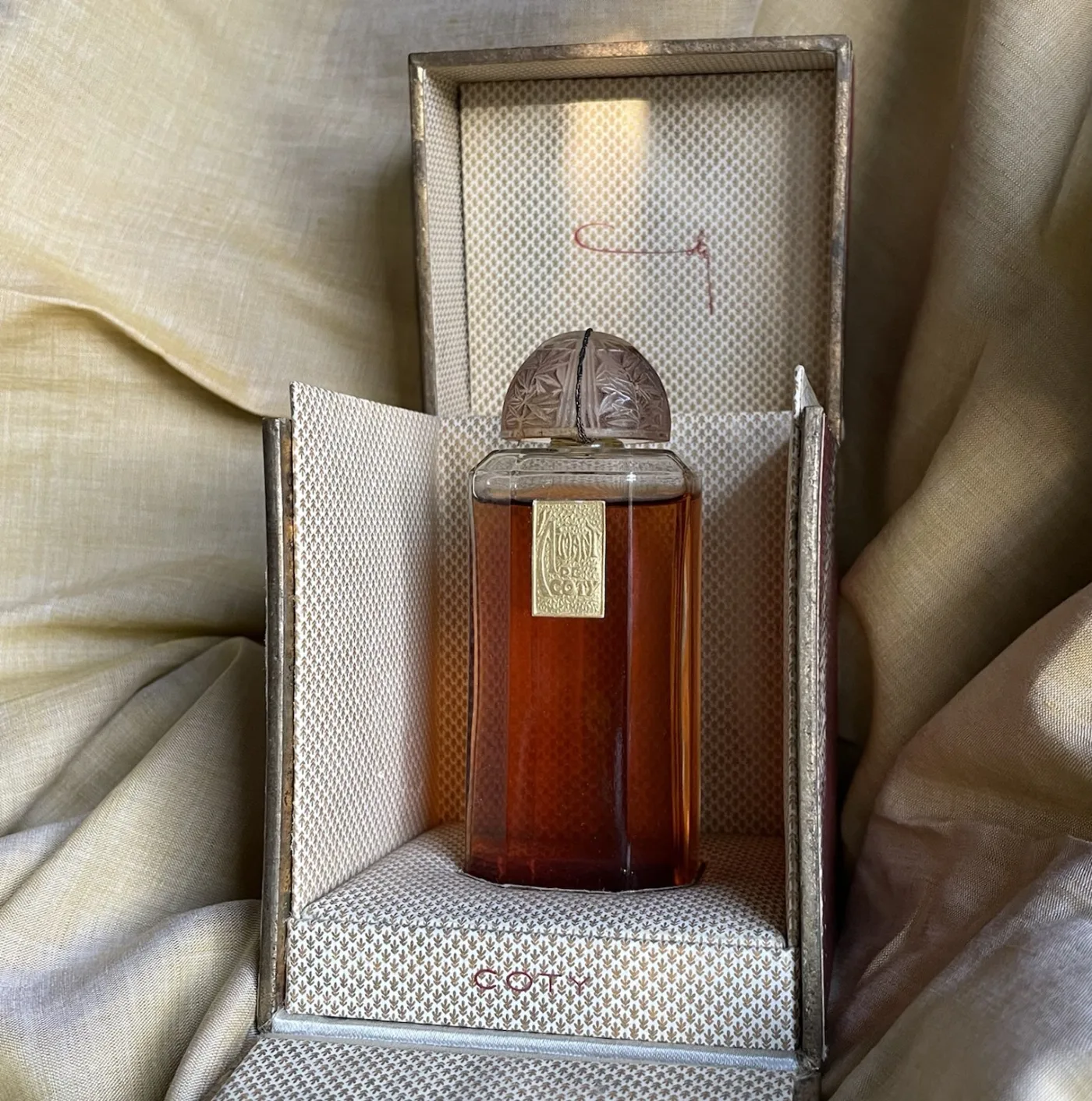 Amber-filled rectangular glass perfume bottle with a frosted moulded stopper and a gold paper label, standing inside its opened cream fabric-covered presentation box printed COTY