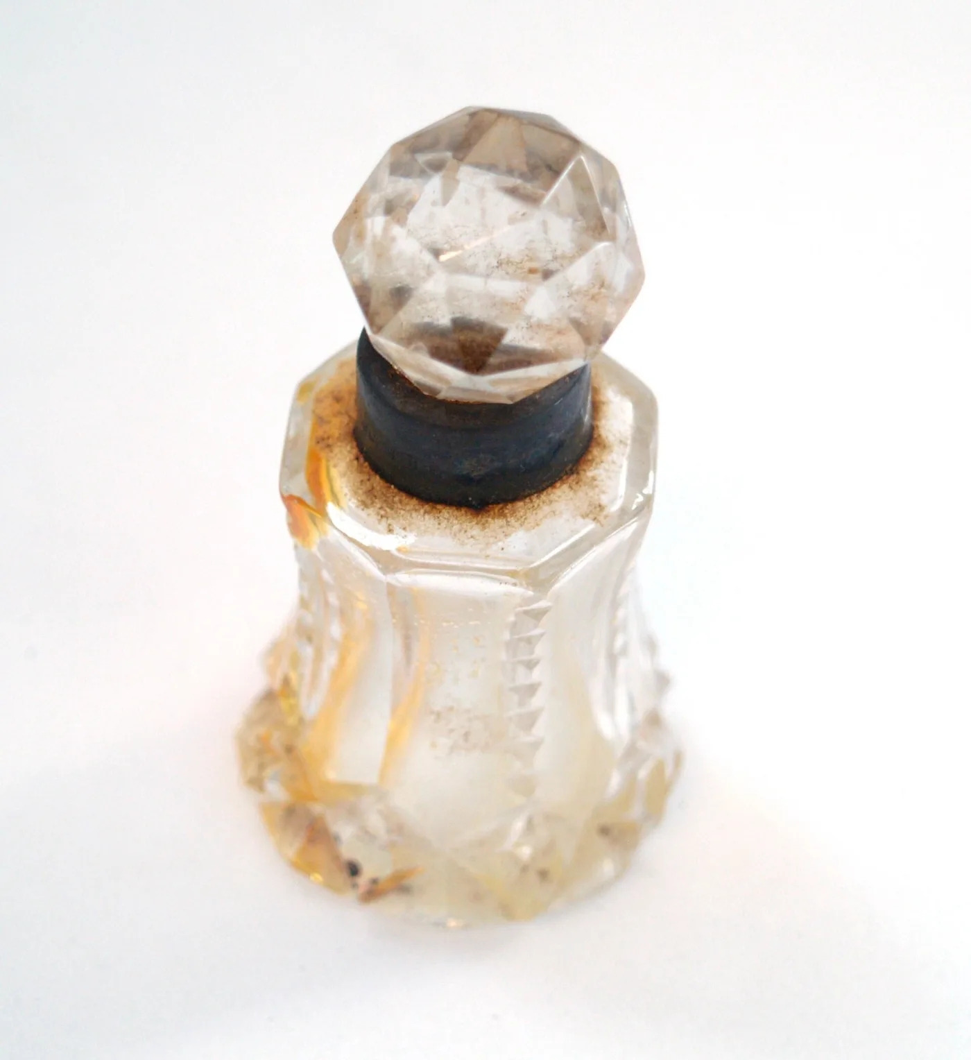 Small clear cut-glass bottle with a broad faceted base and octagonal shoulder, a faceted ball stopper resting in its mouth, and a dark corroded metal collar around the neck under the stopper