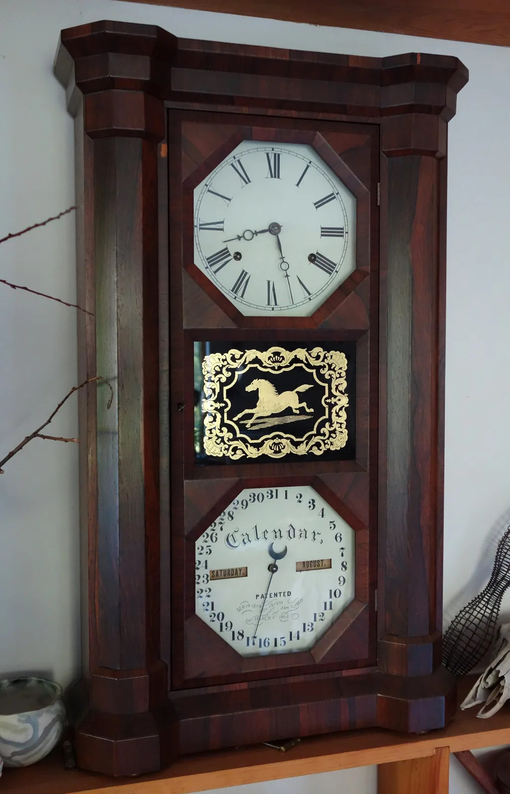 Tall double-dial calendar wall clock in a dark case, a Roman-numeral time dial above and a calendar dial below with day and month apertures and four printed patent dates