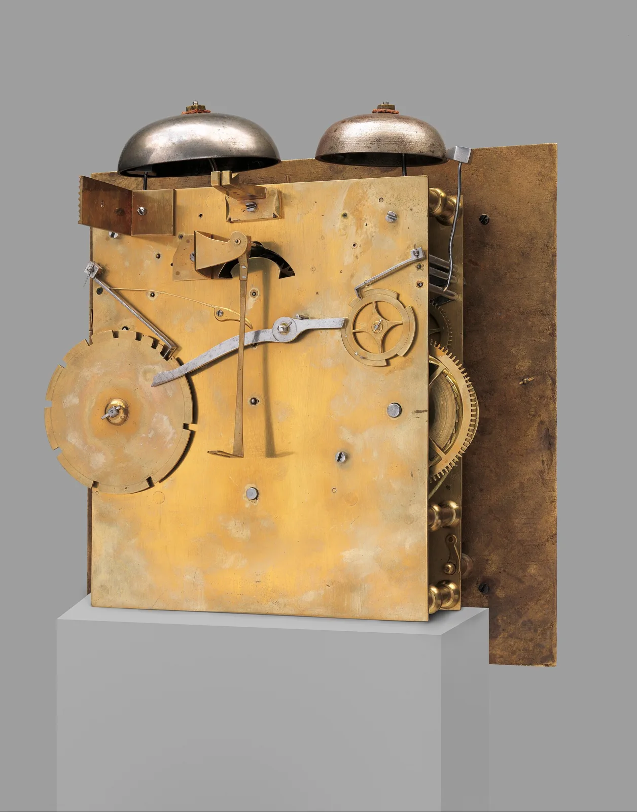 Brass clock movement backplate standing on a plinth, with two domed bells above and a large notched wheel at the lower left