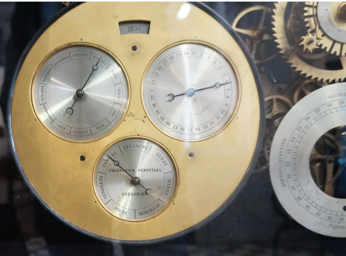 Brass calendar dial with three silvered subsidiary dials for month, date and weekday, and a rectangular aperture above them showing the year 1898