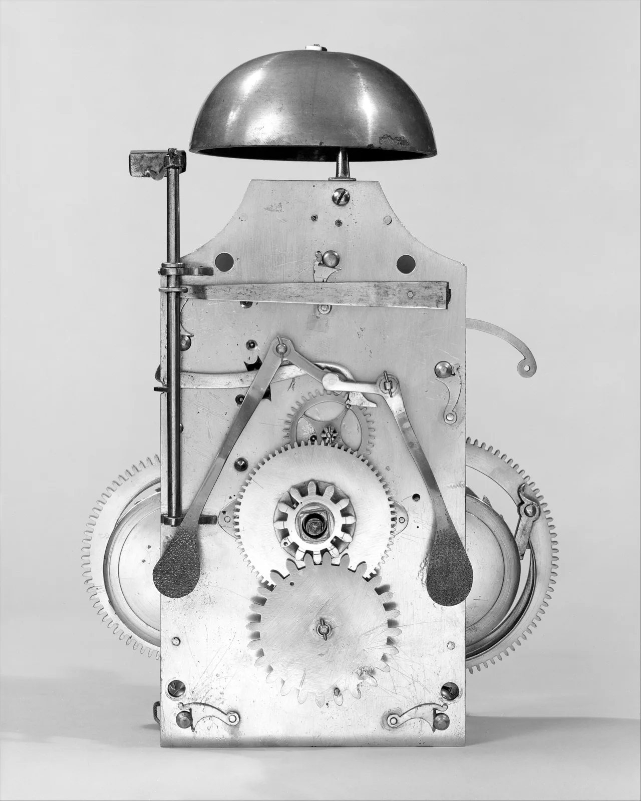 Black and white archival photograph of a narrow clock movement with a domed bell on top and two large toothed wheels projecting at either side