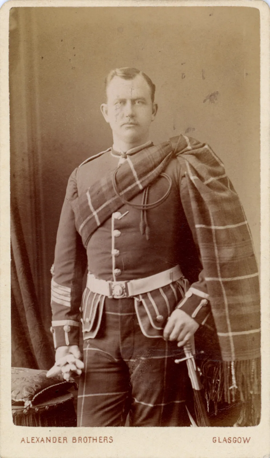 Carte de visite showing a standing soldier in full Highland dress with a tartan kilt and plaid, mounted on a card with the photographer's name printed small along the lower edge