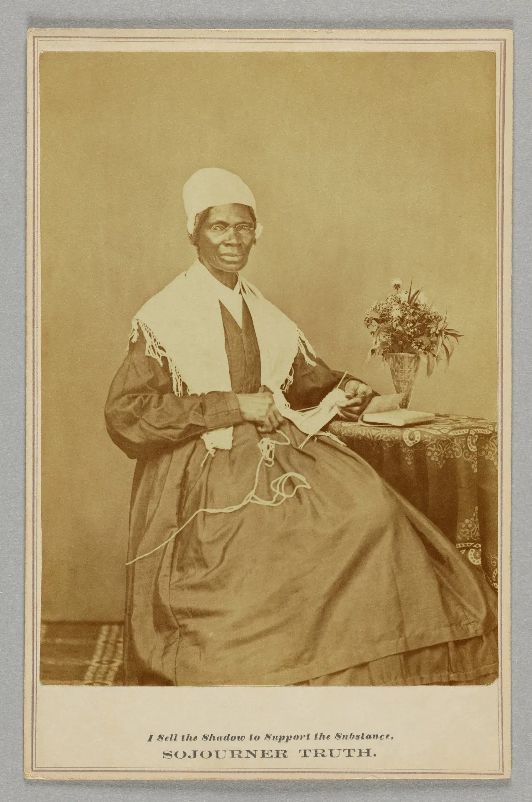 Cabinet card of a seated woman in a shawl and white head covering, her hands resting on a table beside a vase of flowers, mounted on card with a printed caption beneath the image