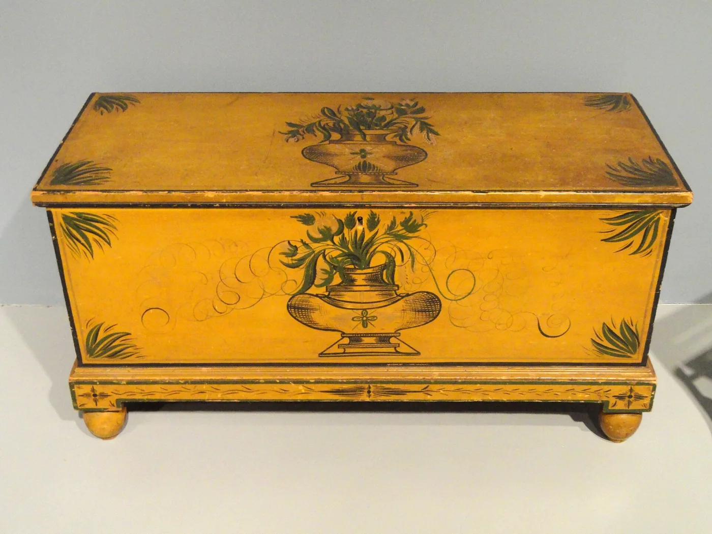 Painted pine blanket chest of 1836 on turned ball feet, its whole surface a bright ochre yellow with a two-handled urn of flowers painted on the front and a smaller one on the lid, both in dark green