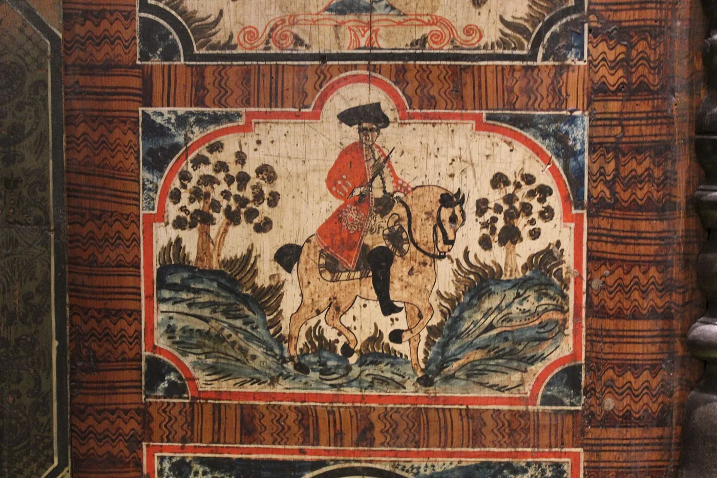 Close view of the central painted panel of the Austrian cupboard, showing a rider in a red coat and tricorn hat on a horse among stylised bushes, inside a scalloped red and white cartouche framed by zigzag combed graining