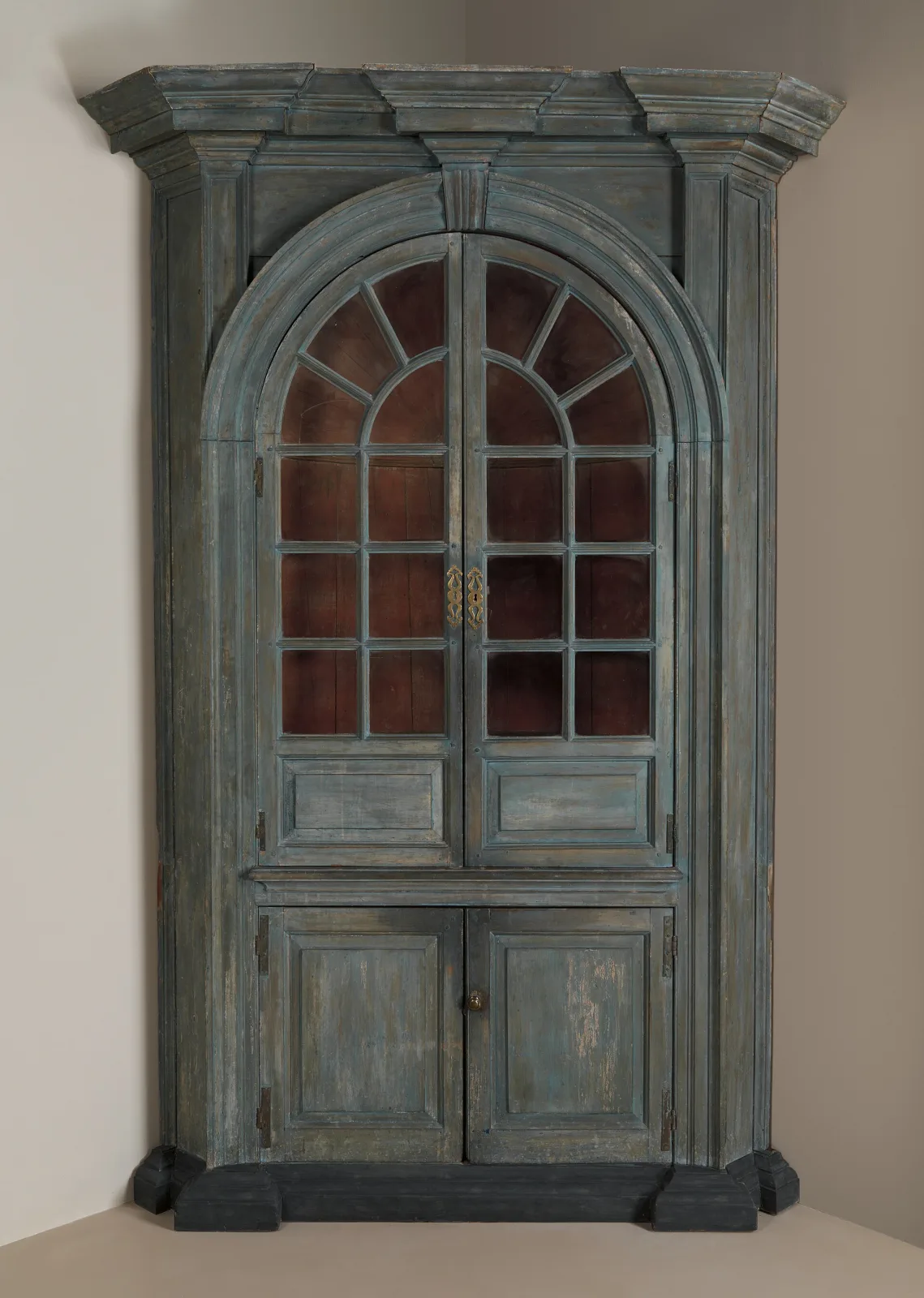 Tall built-in corner cupboard from a Pennsylvania house, with an arched glazed upper section holding shelves, panelled doors below and a broken pediment top, painted a pale colour