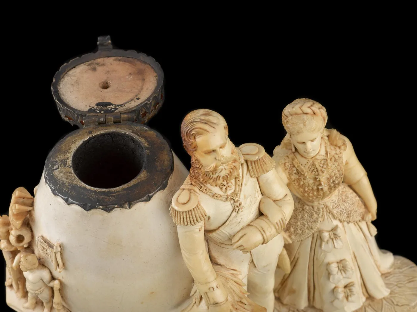 Close view of a meerschaum pipe bowl with a hinged metal rim mount and lid, beside carved figures of a bearded officer and a woman