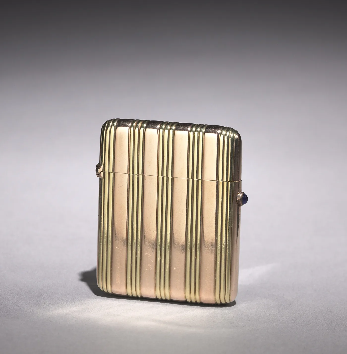 Plain gold vesta case with vertical reeded bands and a blue cabochon sapphire set in the thumbpiece