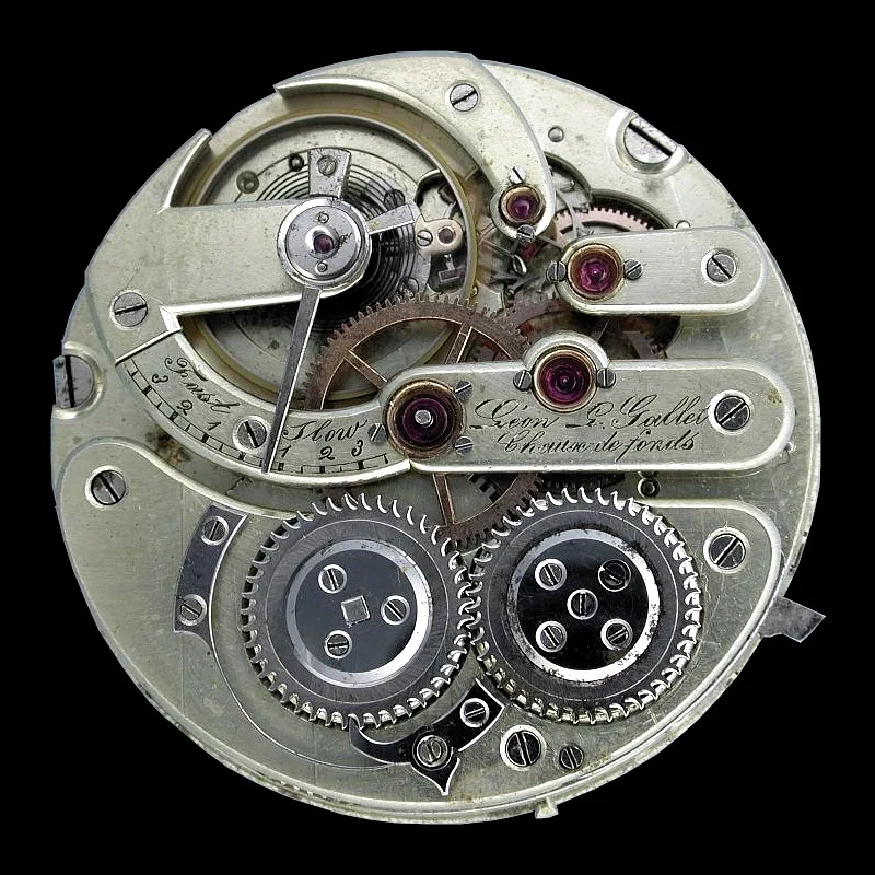 Circular nickel watch movement seen from the back, its plates cut away to show the going train, with several red jewel bearings set into the plate and an engraved maker's signature across it
