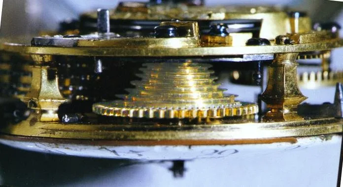 Close side view inside a watch movement showing a small stepped brass cone wound with fine chain, standing between the plates beside a toothed wheel