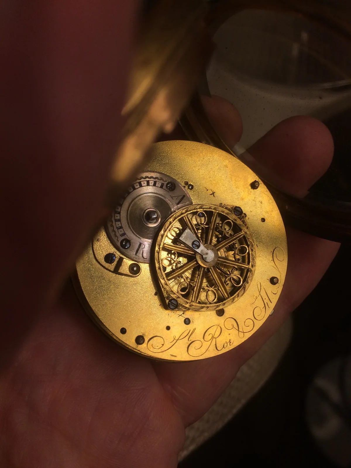 Gilt watch movement held in a hand, its back plate engraved all over with foliate scrollwork around a pierced balance cock, with a signature engraved along the lower edge
