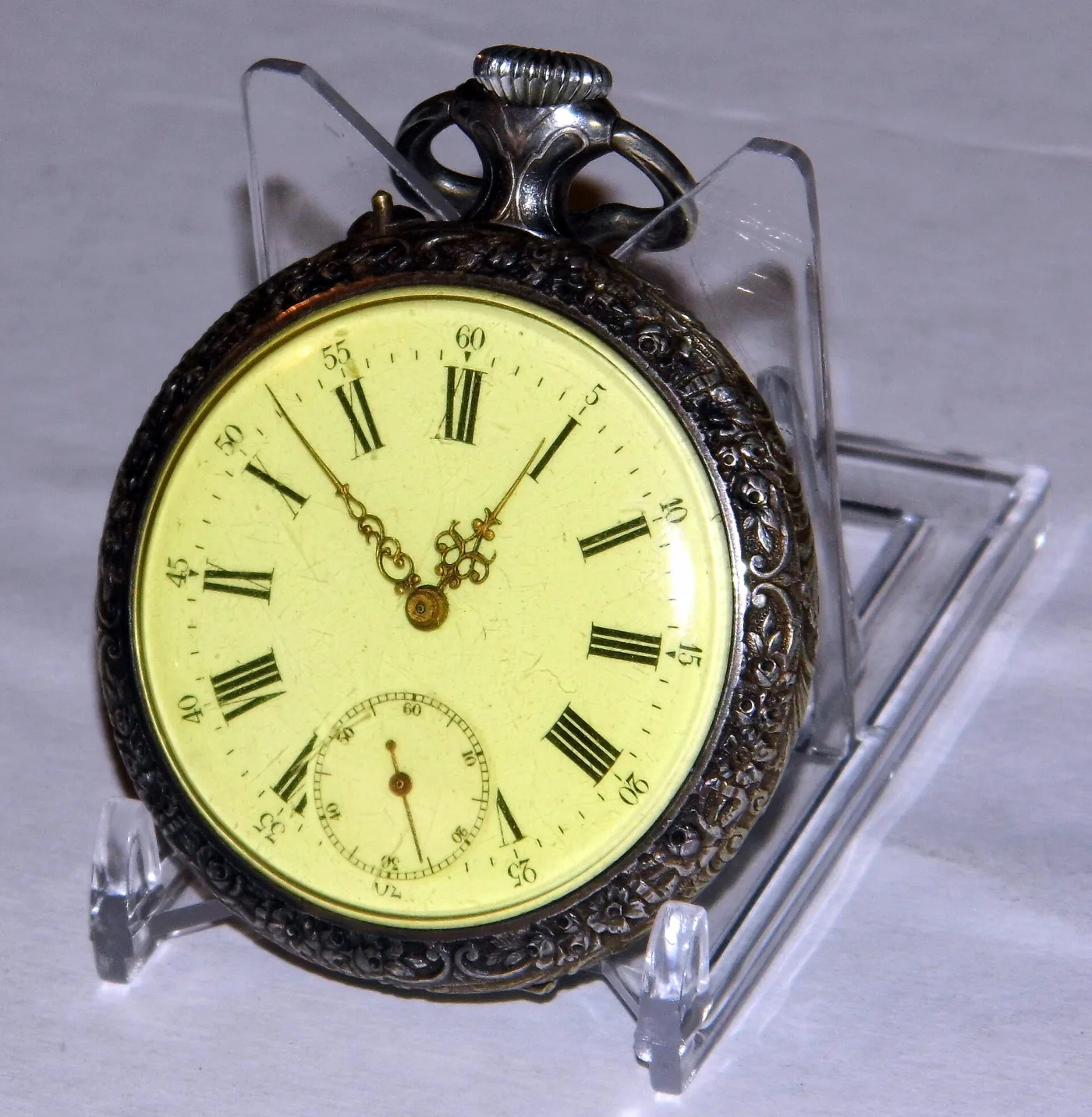 Large pocket watch standing in a clear display support, with a cream enamel dial carrying bold black Roman numerals, an outer minute track, a subsidiary seconds dial and ornate gilt hands