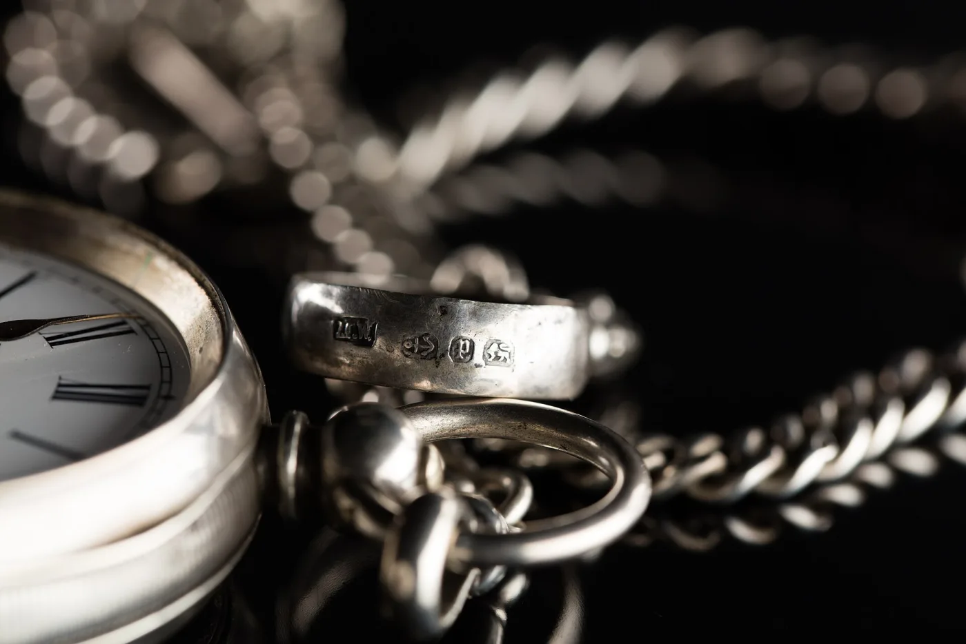 Close view of the join between a pocket watch's silver case and its chain, showing a row of small struck hallmarks punched into the metal at the pendant