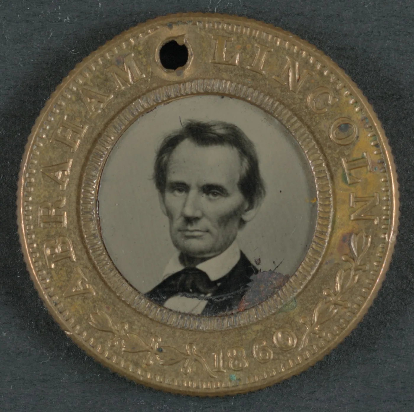 Small 1860 campaign badge with a brass rim stamped ABRAHAM LINCOLN and the date 1860 around a circular tintype portrait of Lincoln, a suspension hole punched through the top of the rim