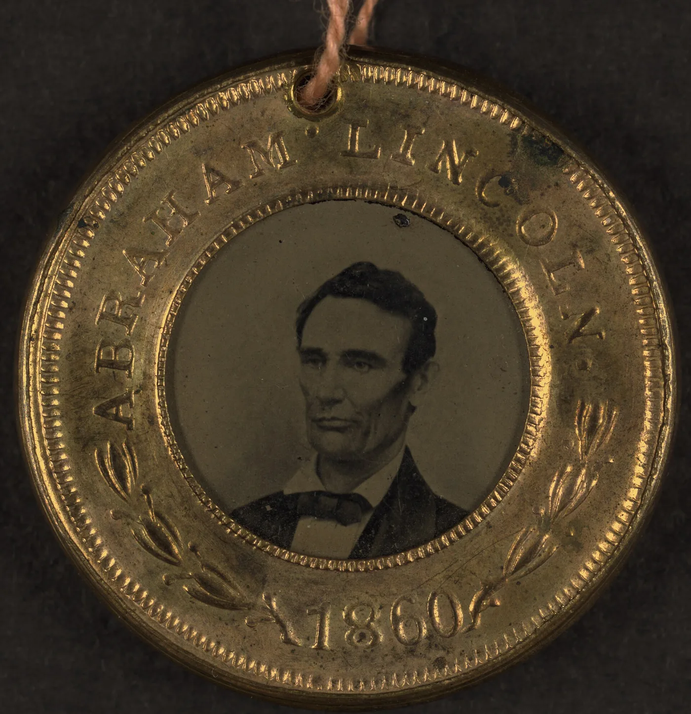 Close view of an 1860 tintype campaign badge, showing the mottled and clouded surface of the tintype, wear to the gilding of the stamped rim and shallow scratches across the image