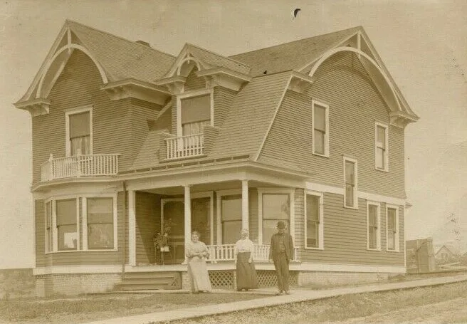 Sepia real photo postcard of a two-storey clapboard house with a gambrel roof, a man and two women standing on the path in front of it