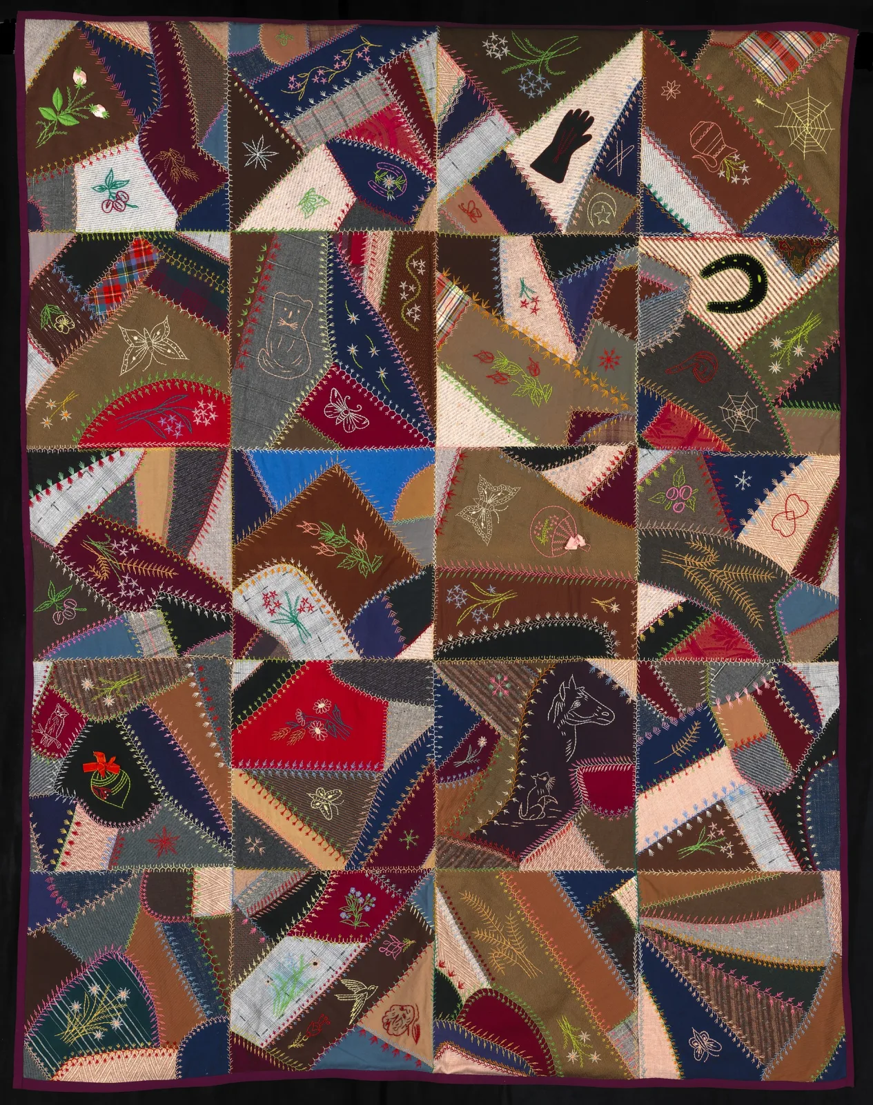 Quilt of irregular angular patches of dark silk, velvet and wool, every seam covered with coloured embroidery and scattered with embroidered motifs including a spider web, a horseshoe, a glove and a horse
