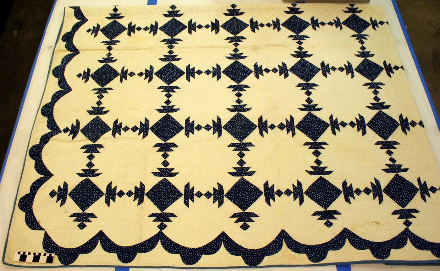 Indigo and cream pieced quilt laid flat on a table for photography with a black-and-white scale bar at one corner, showing a scalloped indigo border and brown staining near one edge
