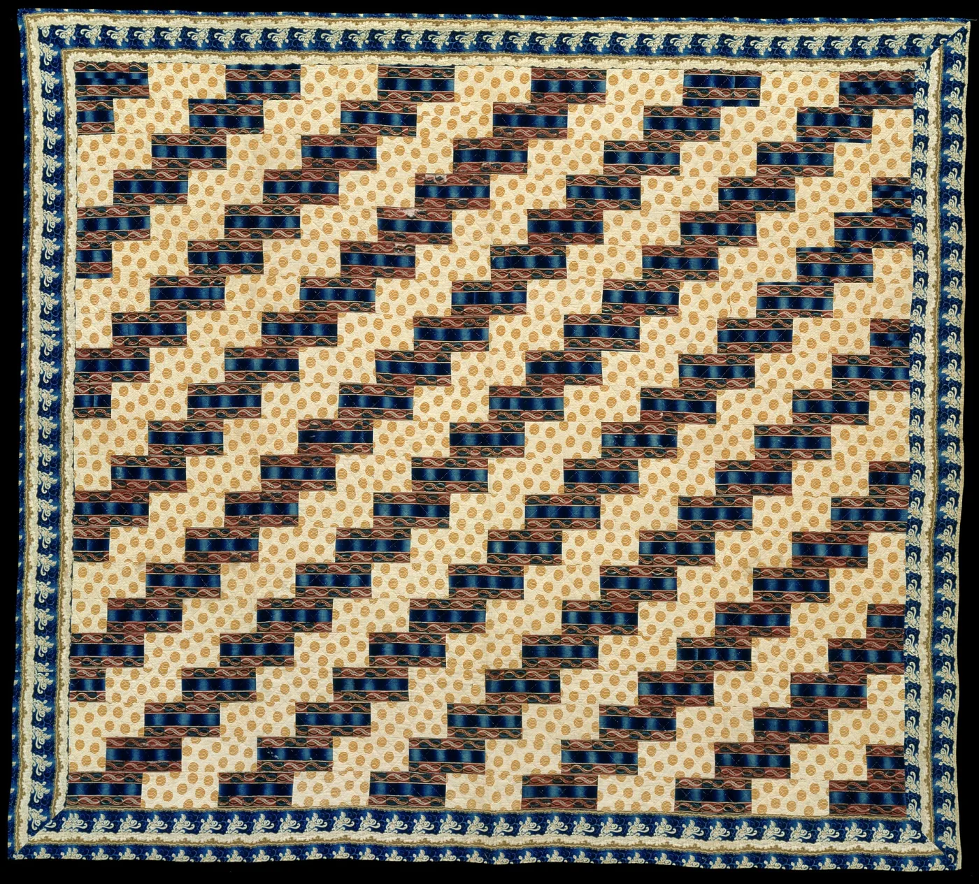 Quilt of small rectangular patches in dark indigo, ochre dots and rust stripes laid in staggered rows like brickwork, inside a deep blue printed border
