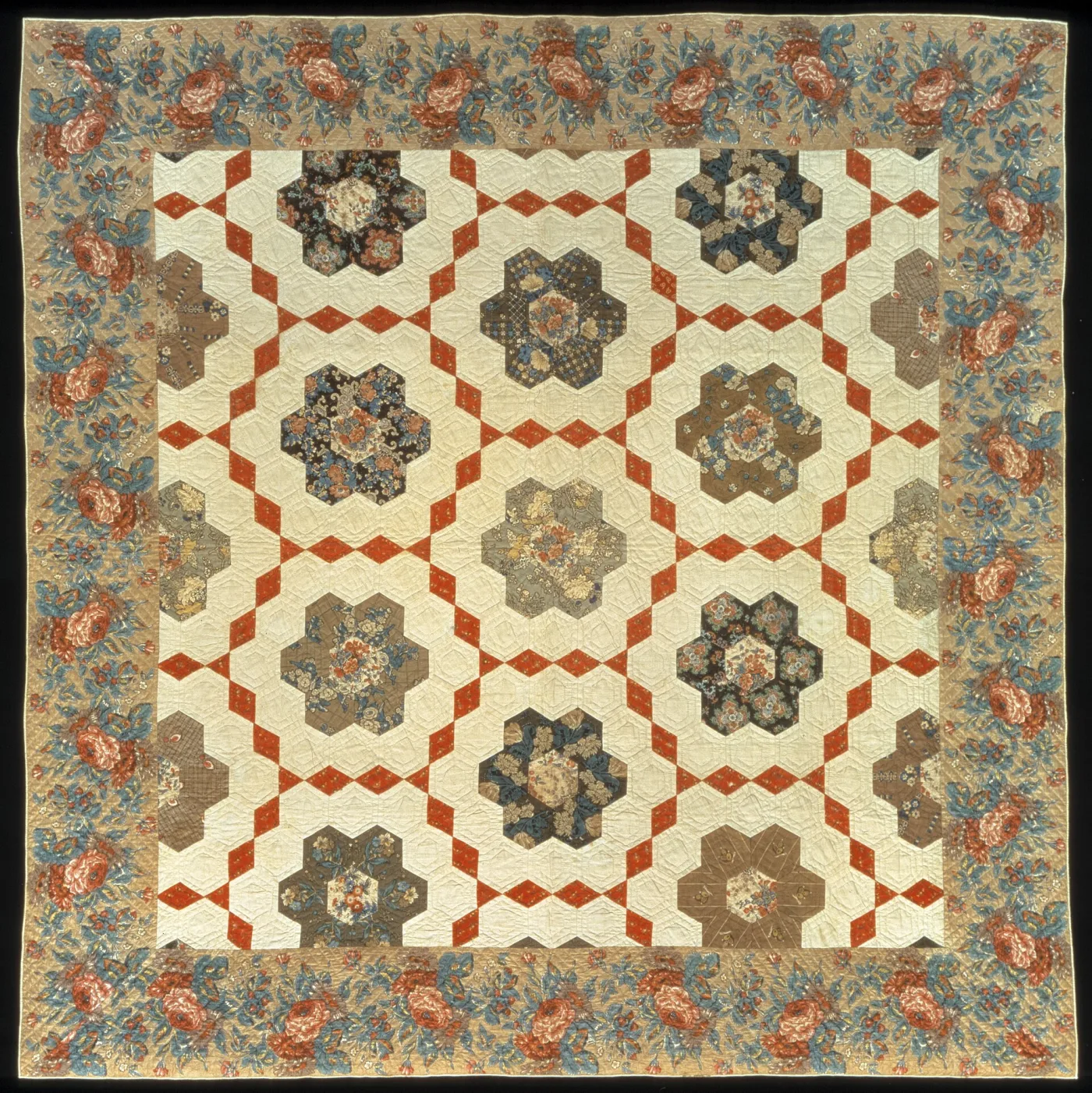 Quilt of six-sided floral chintz rosettes, each built from seven hexagons, set in a white ground crossed by red diamond lattice, inside a wide chintz floral border