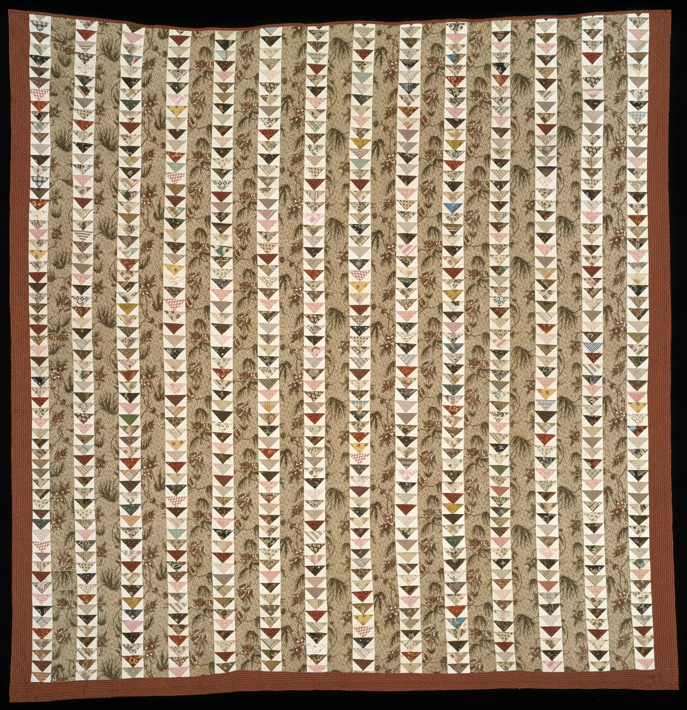 Quilt of vertical strips, each strip a column of small triangles all pointing downward, alternating with strips of printed brown-and-green foliage fabric