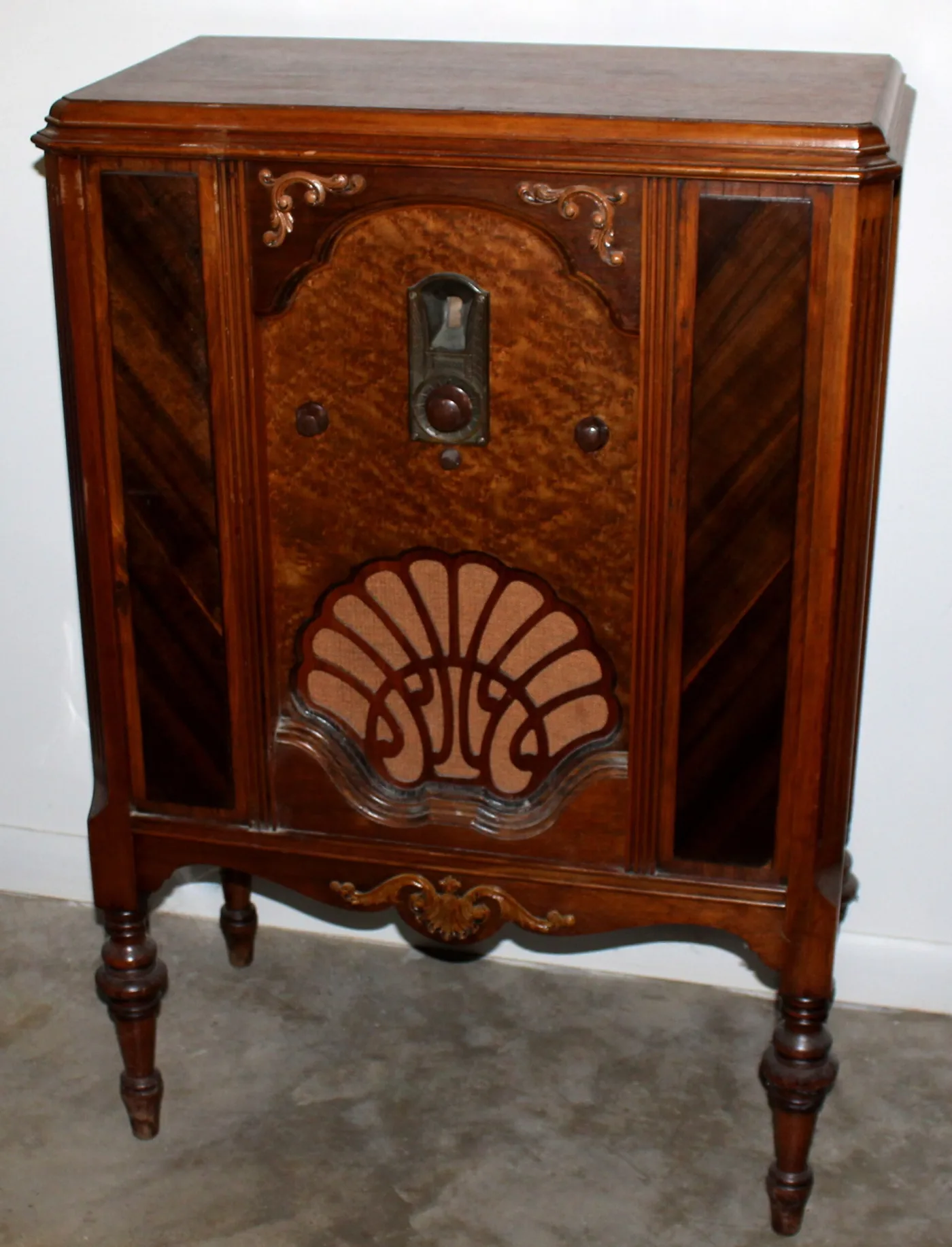 Waist-high wooden radio cabinet on turned legs, with a carved shell-shaped speaker grille, a small arched dial and applied scrollwork