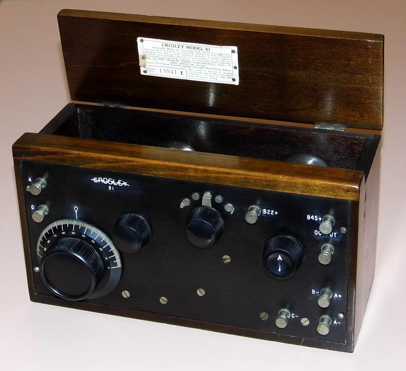 Open wooden box radio with a black panel, one large tuning dial, three knobs and rows of terminal posts, with a maker's label inside the hinged lid