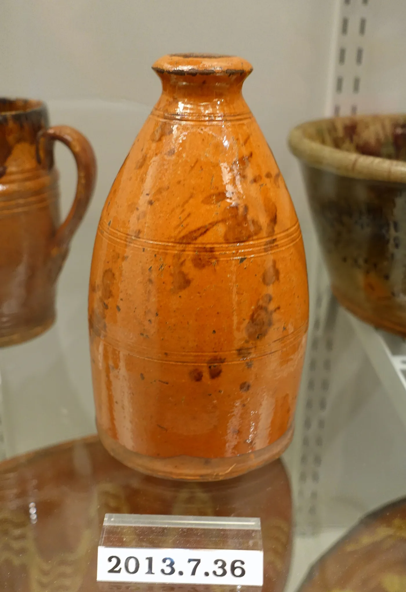 Tall orange-glazed redware bottle with scattered dark brown blotches and two tooled bands, on a museum shelf