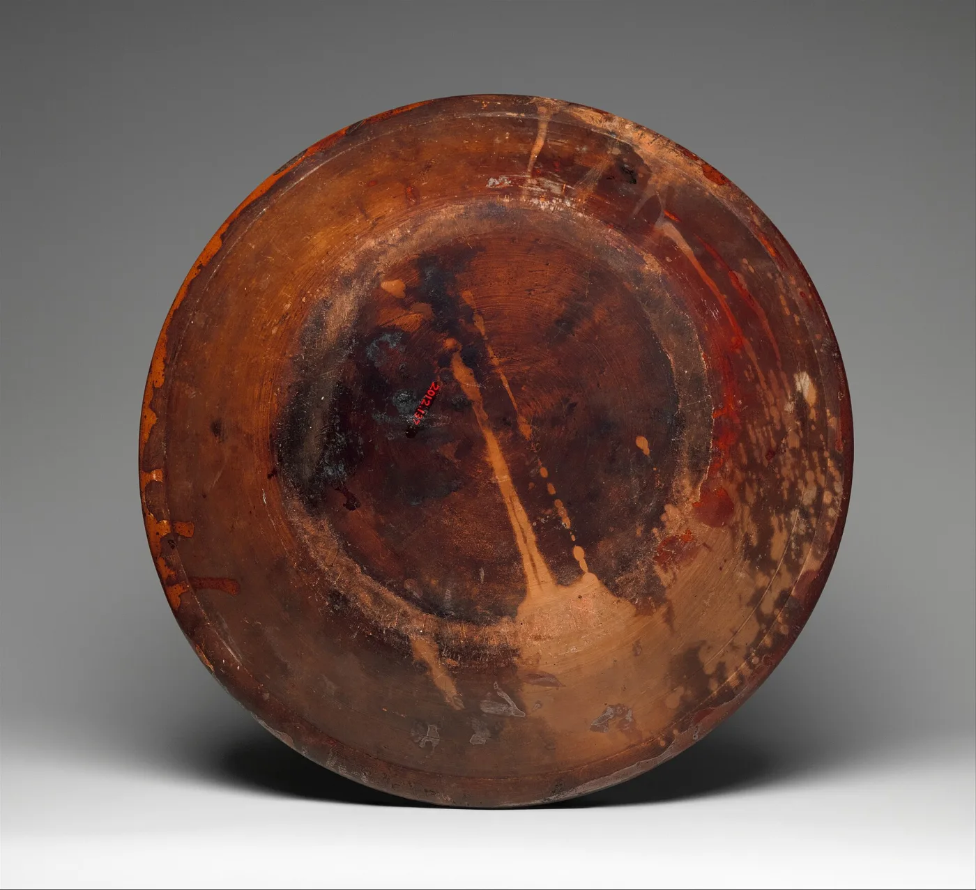 Underside of a redware dish, flat and thinly glazed, with dark kiln clouding, pale stacking scars and a small painted accession number