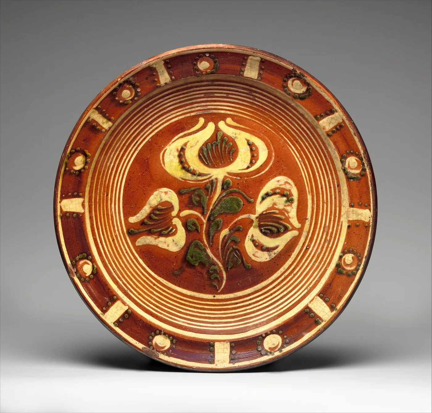 Round red earthenware dish decorated with a cream and green tulip spray inside concentric slip rings, with a barred and dotted border