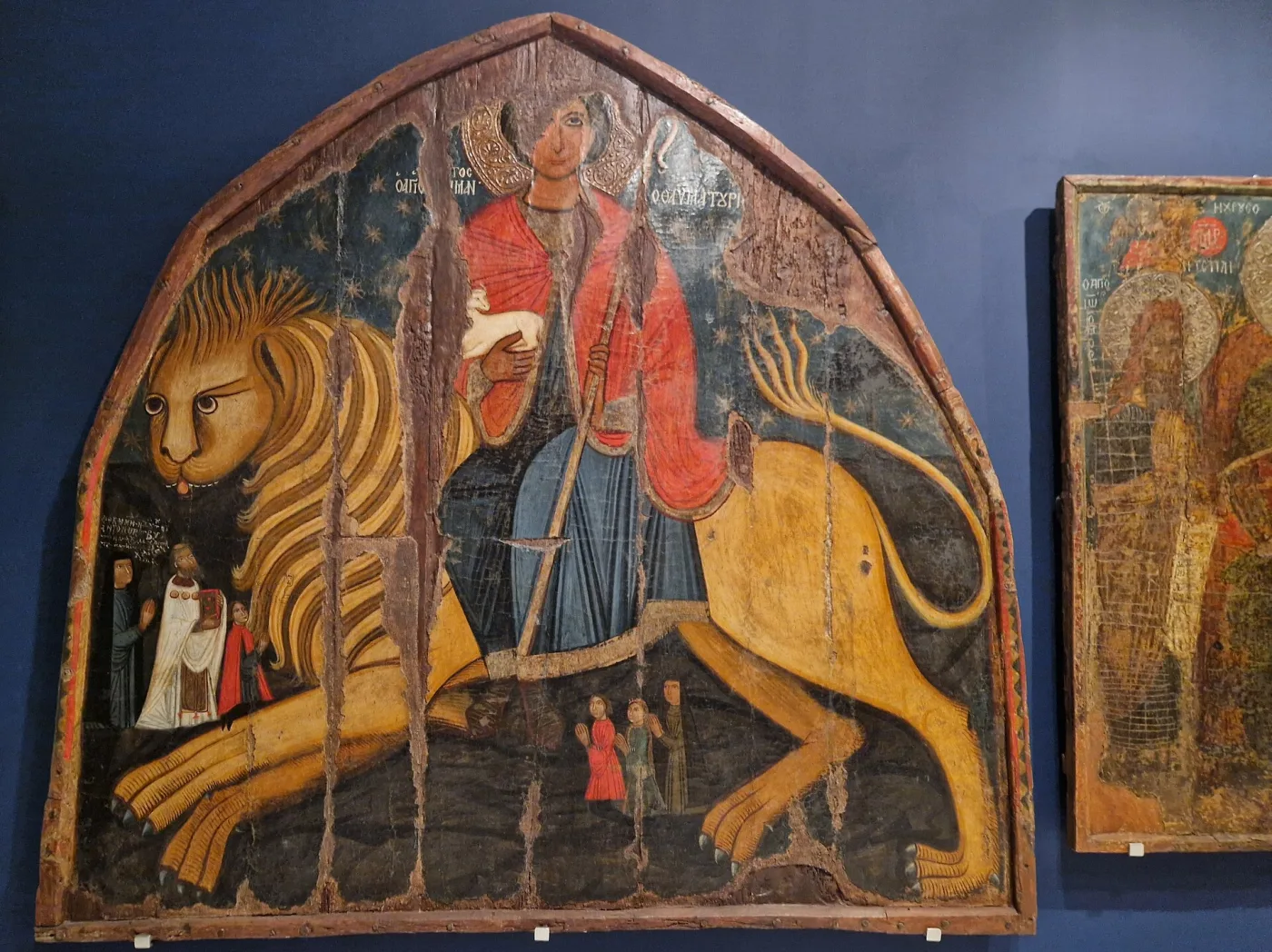 Arched wooden icon panel showing a haloed saint in armour and a red cloak riding a large golden lion, with small figures at the lower left, displayed in a museum