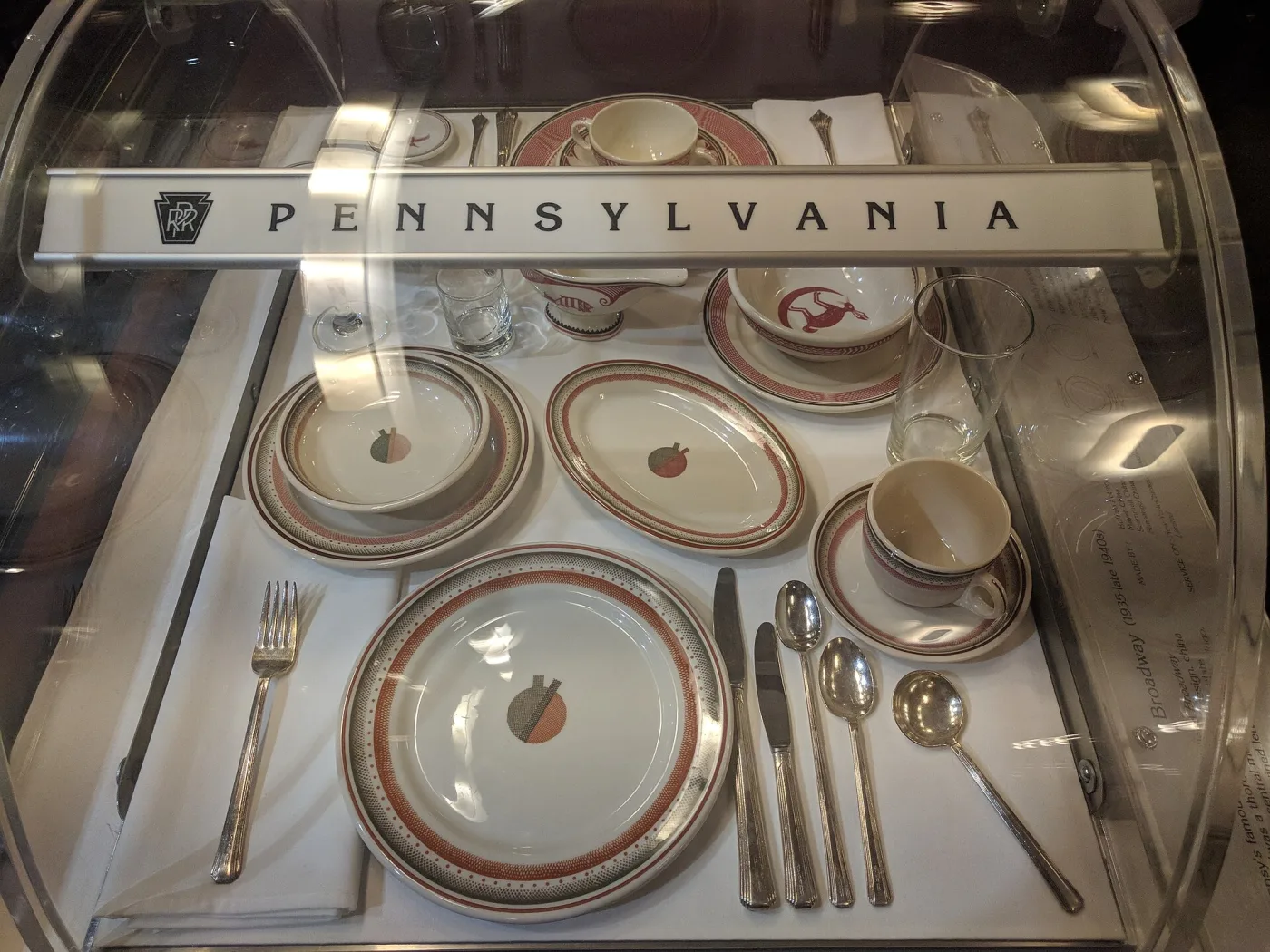 Museum case of Pennsylvania Railroad dining car china with red and grey banded rims and a small stylised motif at the centre of each plate