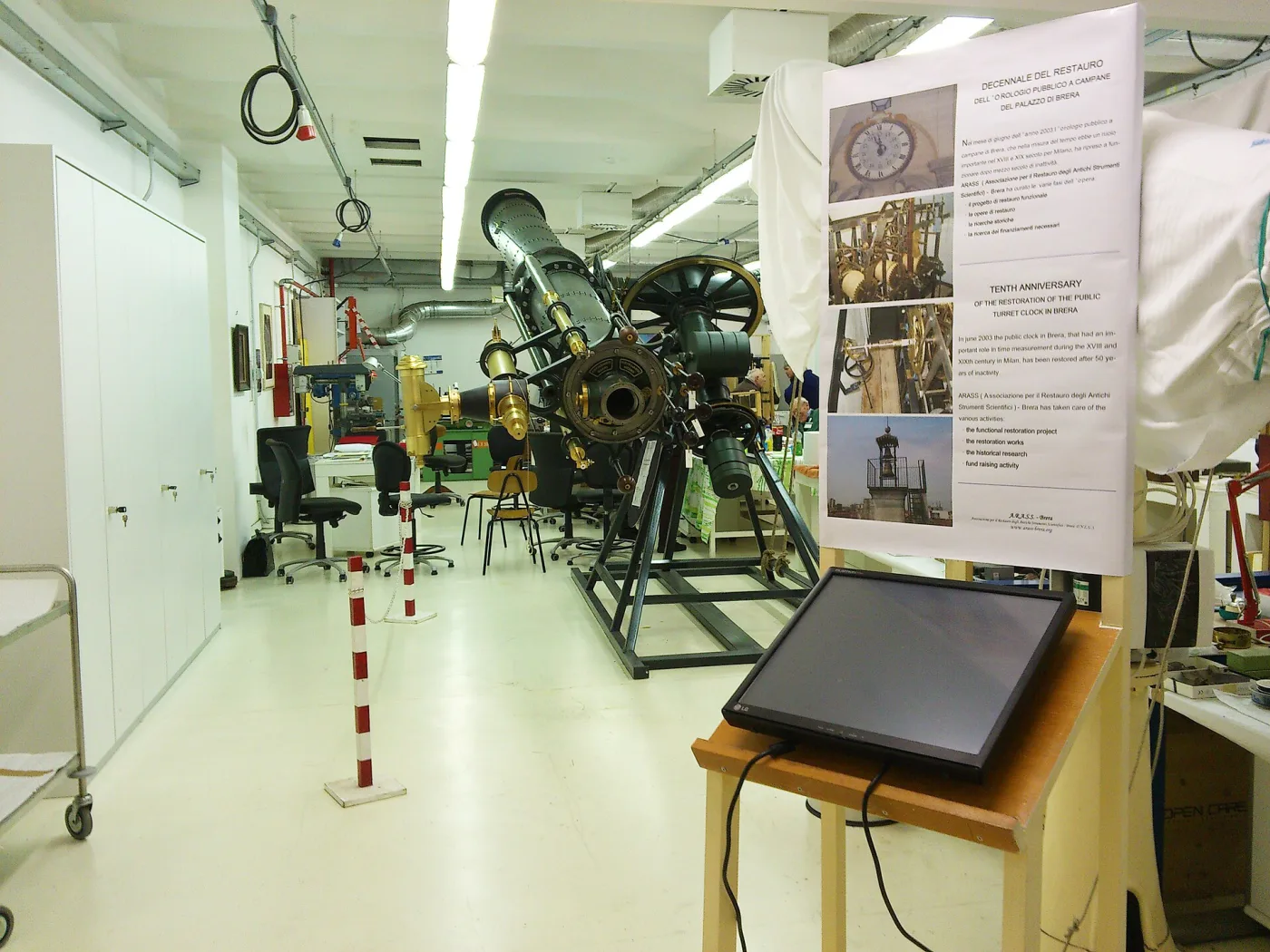 A long workshop containing a large brass and iron equatorial telescope on a cast frame, beside a bilingual display panel about the restoration of a public turret clock