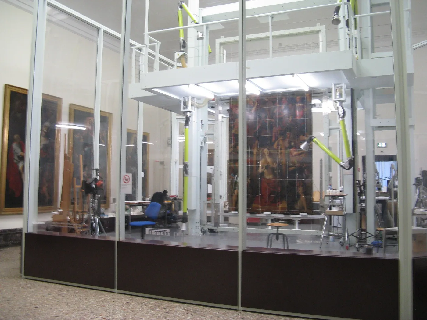 A glass-walled conservation laboratory inside a gallery, containing a large painting on a gridded support, extraction arms overhead, easels and further paintings stacked against the wall