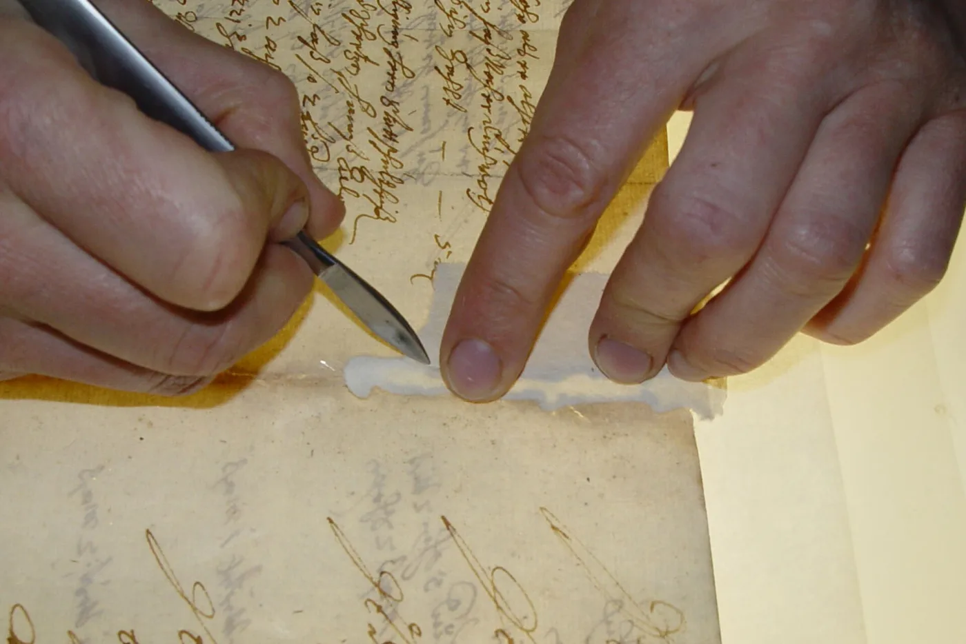 Close view of two hands repairing a tear in a handwritten document, one holding a scalpel and the other pressing down a strip of pale Japanese tissue