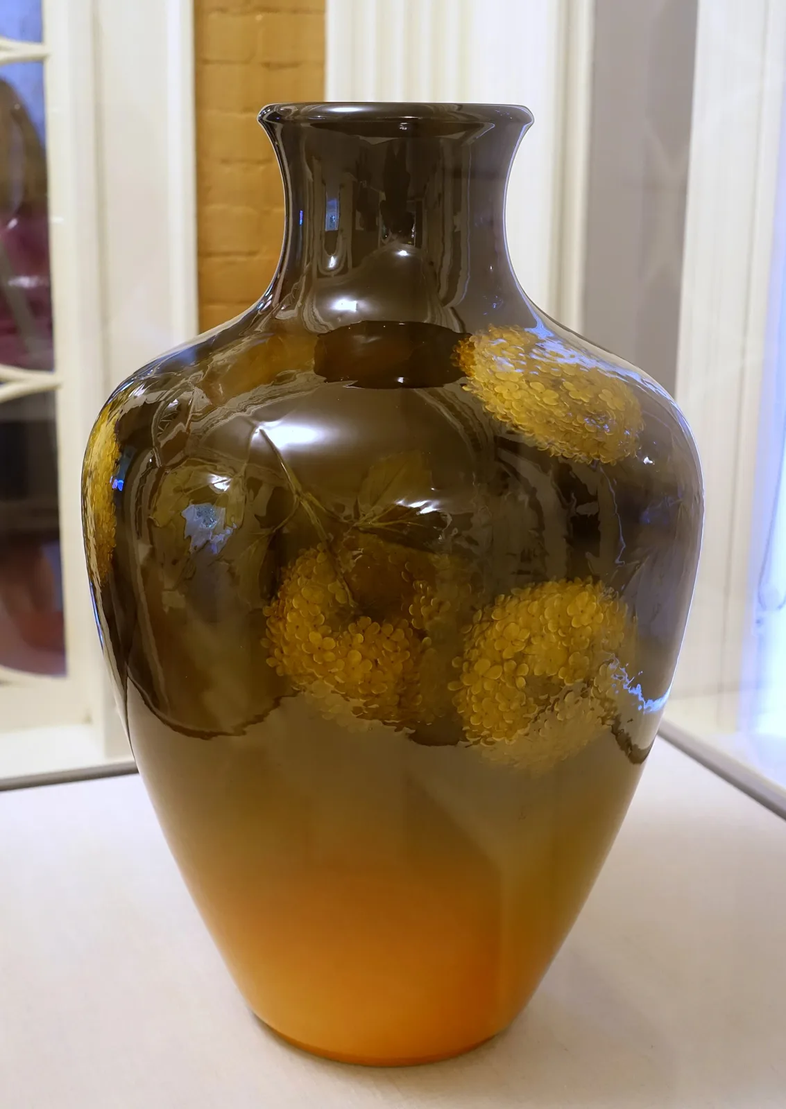 Swelling ceramic vase shading from deep brown at the shoulder to warm orange-yellow at the base, slip-painted with yellow chrysanthemums and green leaves under a thick glossy amber glaze