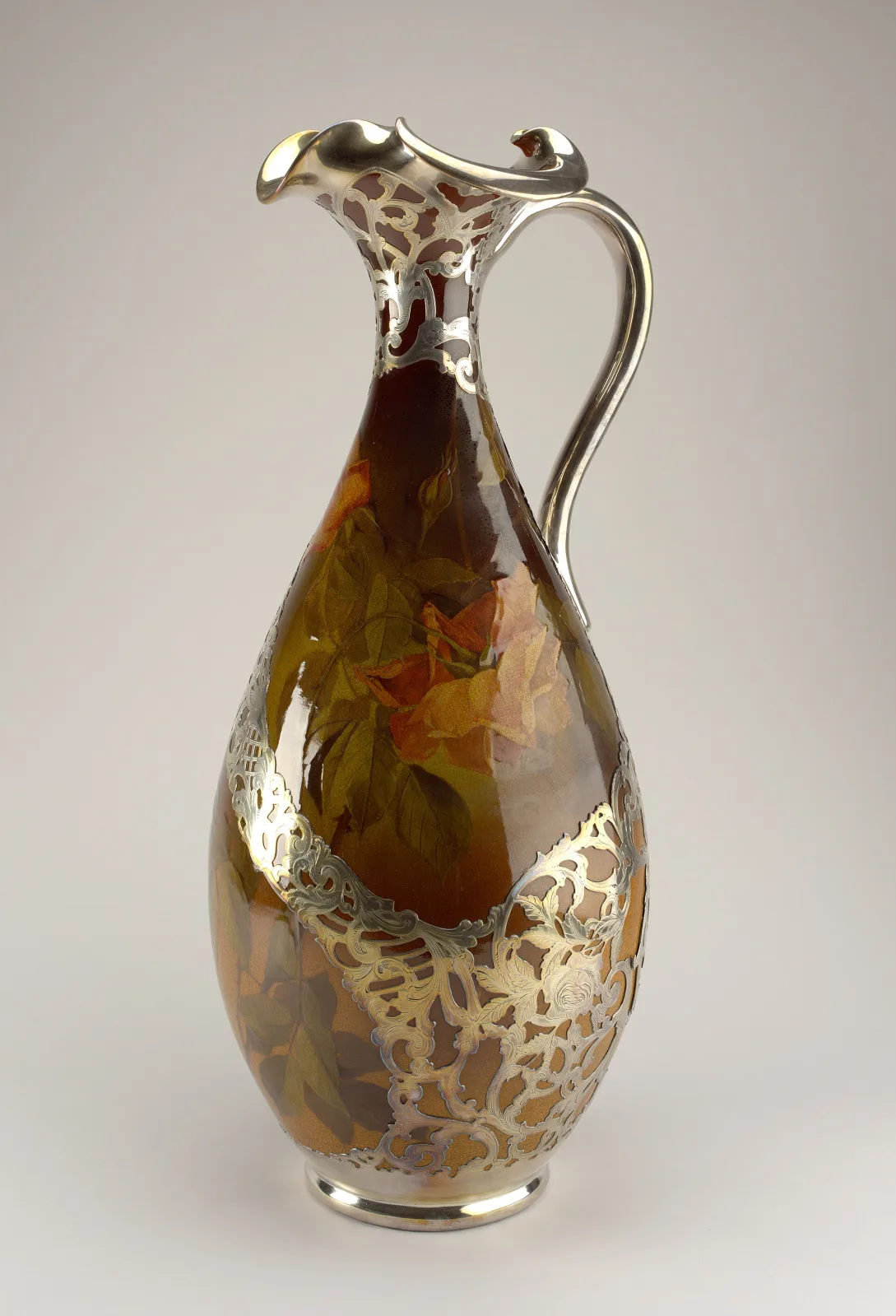 Slender ceramic ewer in dark glossy brown and amber, slip-painted with autumn foliage, its neck, spout, handle and foot encased in pierced and chased silver mounts