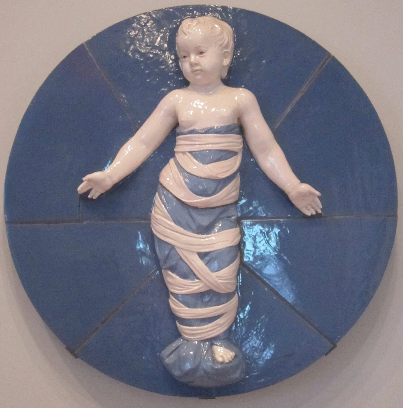 Circular ceramic plaque built from shaped tiles, showing a swaddled infant modelled in high relief in cream and pink against a deep blue ground