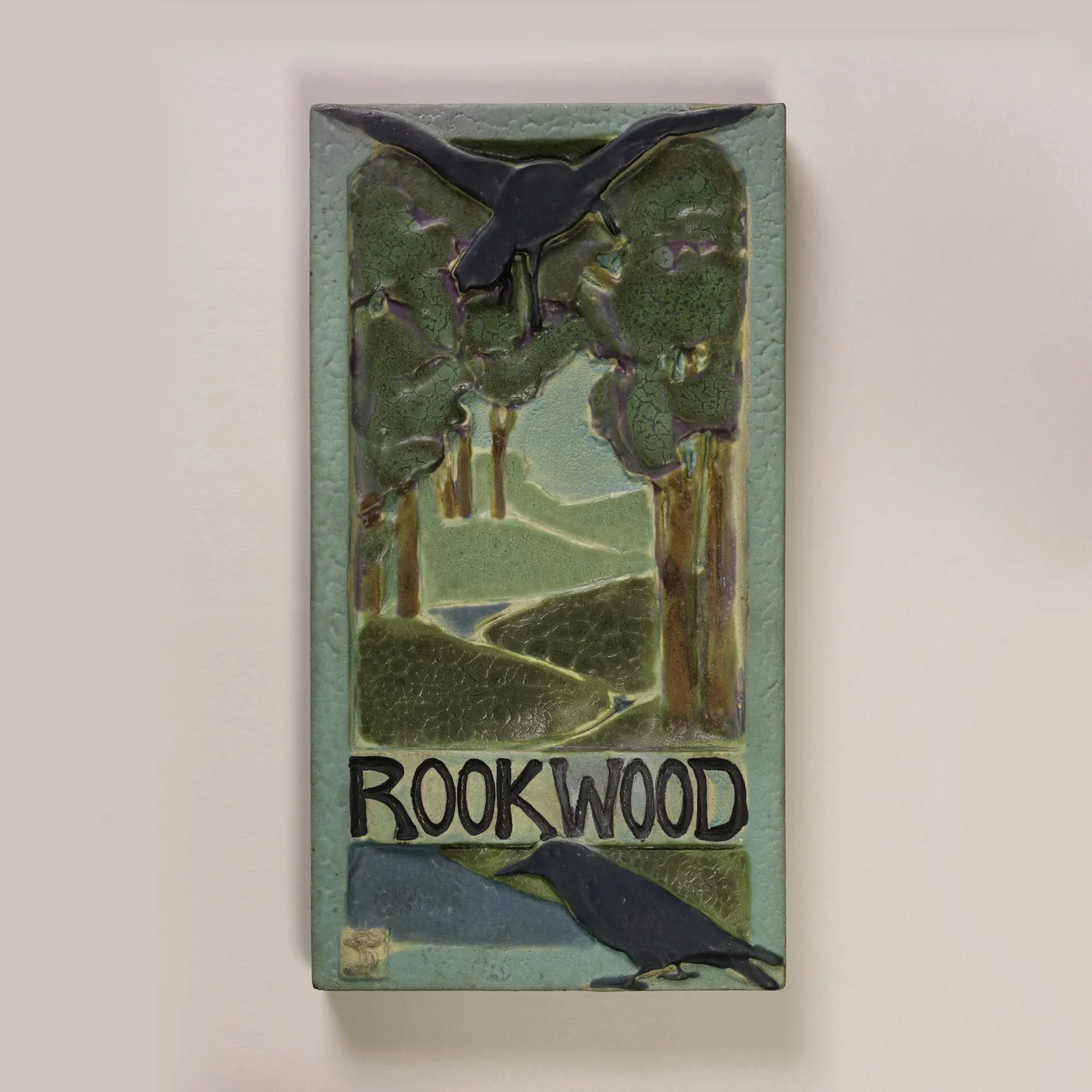 Tall rectangular tile panel moulded in low relief with two dark rooks perched among trees above a green landscape, the name ROOKWOOD lettered in raised capitals across the lower third