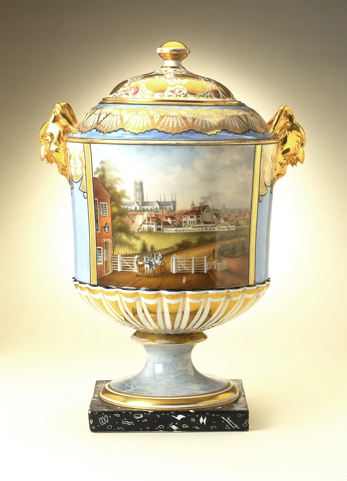 Covered porcelain vase on a black plinth painted with a panoramic view of a town with a cathedral, factory buildings and a horse and carriage, with gilded lion-mask handles