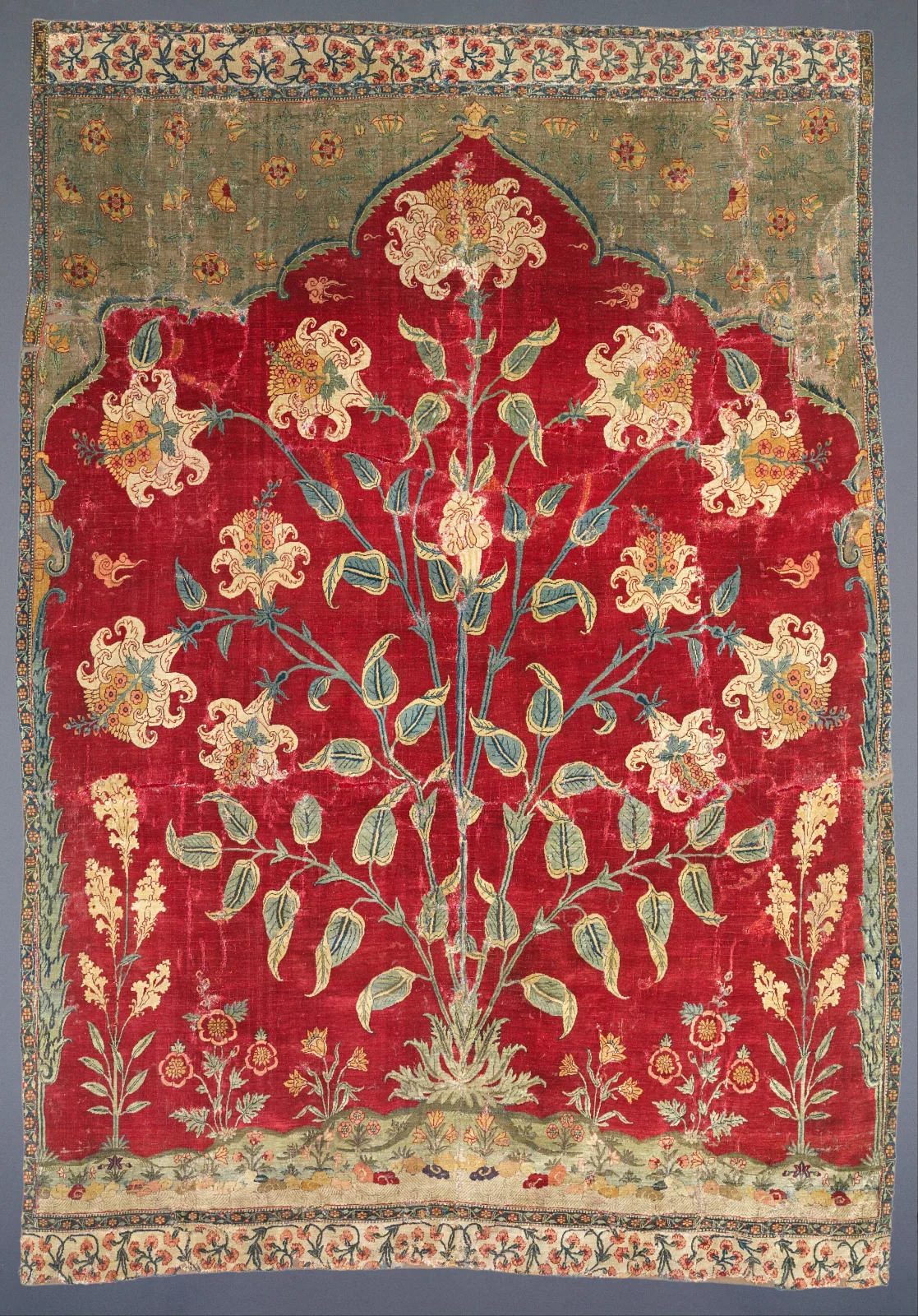 Fragment of an Indian saf carpet of 1600 to 1650 showing one red niche containing a large flowering plant with white and pink blooms, the surrounding ground worn to pale green