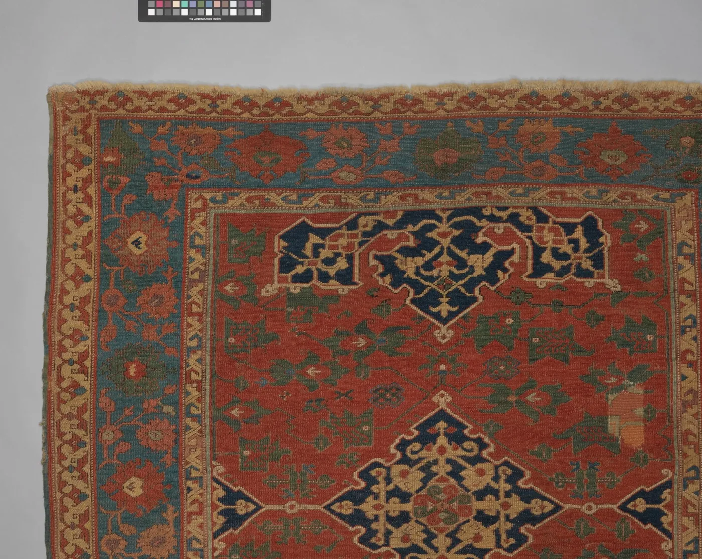 Corner and part of the field of a Star Ushak carpet, probably seventeenth century, showing a large dark blue star medallion on a red ground with a blue and ochre border, photographed beside a colour reference strip